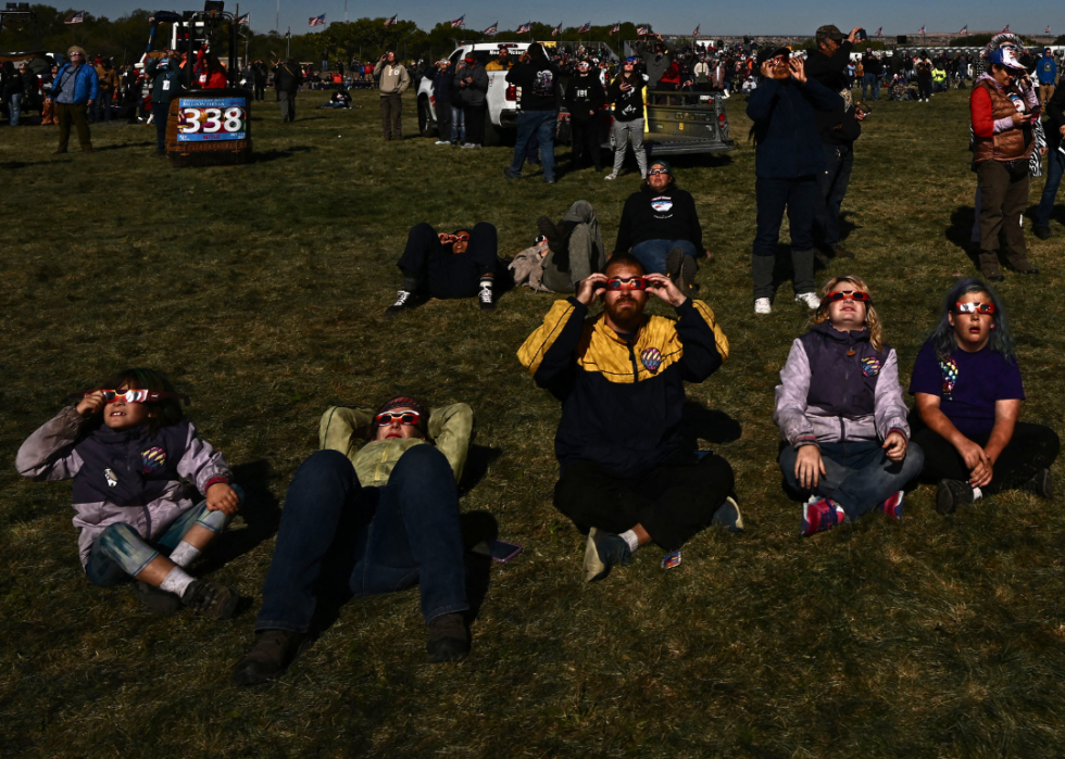 A crowd of people sitting on the grass in large field looking up into the sky wearing cardboard sunglasses.